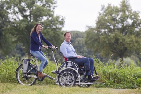 Een dame met lang lichtbruin haar, gekleed in blauw vest en lichte spijkerbroek zit op de rolstoelfiets. Op het plateau zit een witte man gekleed in een licht overhemd en een donkere spijkerbroek in zijn rolstoel. Het dup kijkt vriendelijk. Zij fietsen do