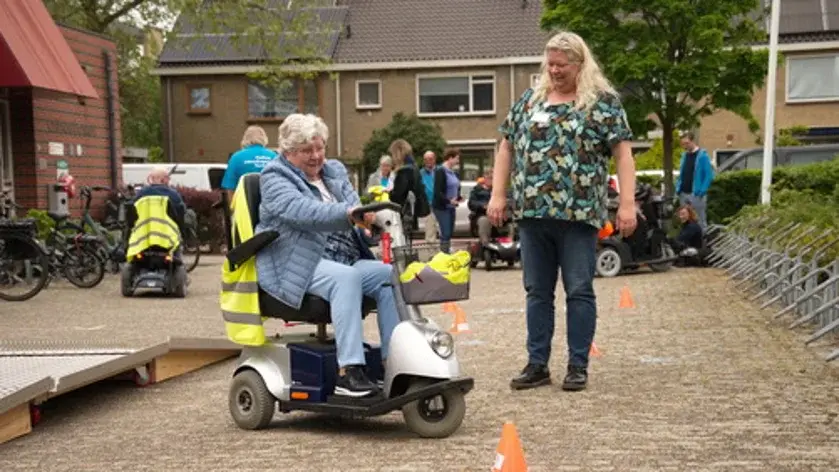 Een blonde dame, gekleed in kleurig shirt en een spijkerbroek, kijkt toe hoe een oudere dame, gekleed in een lichtblauw jack en een lichte spijkerbroek, een scootmobiel bestuurt.
