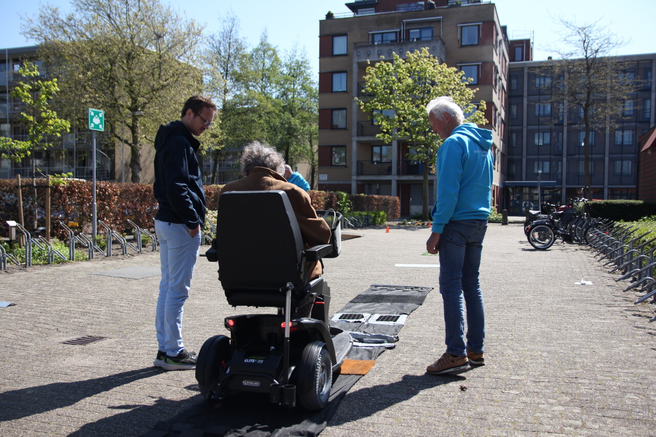 Achteraanzicht van een vrouw op een scootmobiel. Zij rijdt over een parcours, vergezeld door twee deskundigen