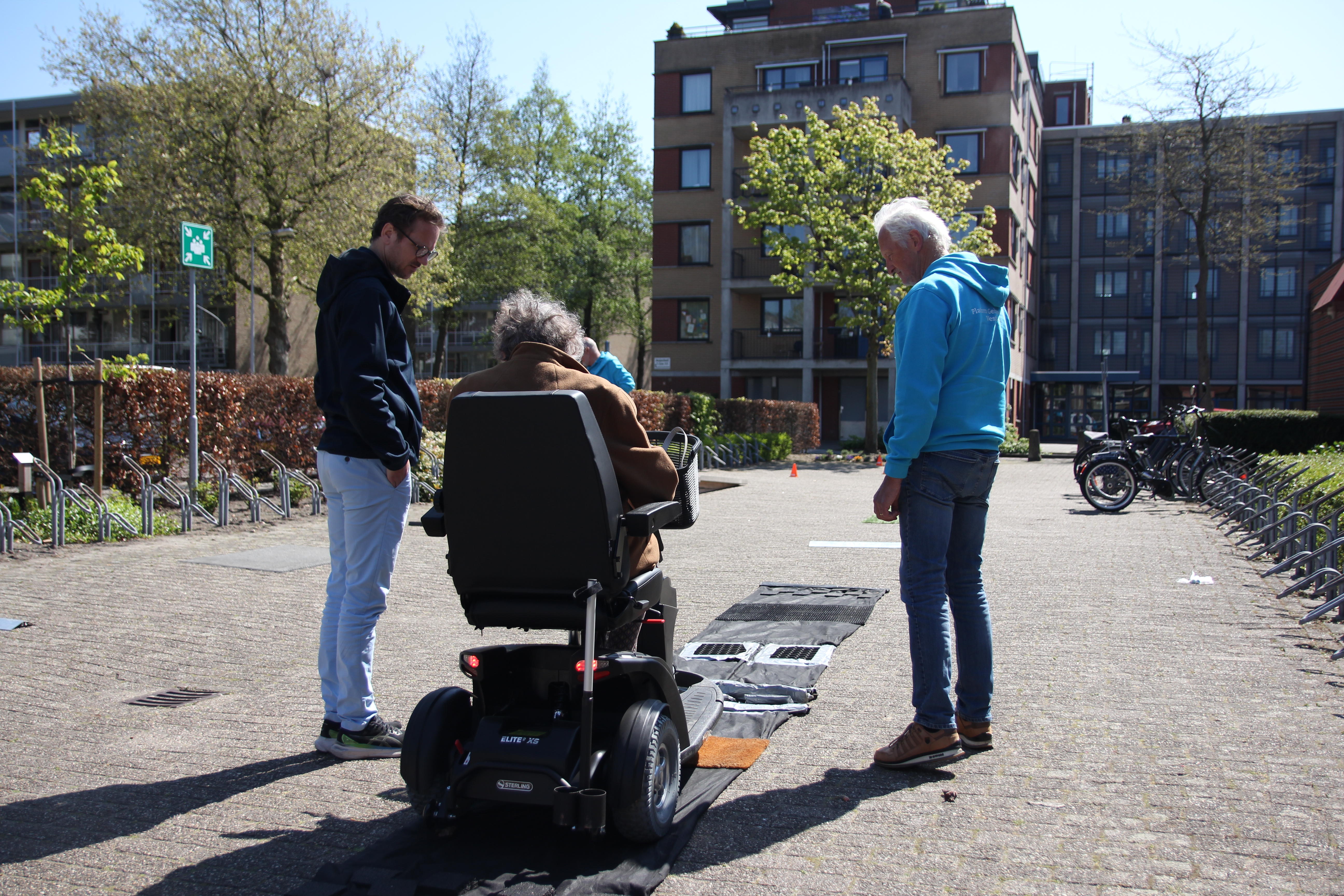 Achteraanzicht van een vrouw op een scootmobiel. Zij rijdt over een parcours, vergezeld door twee deskundigen